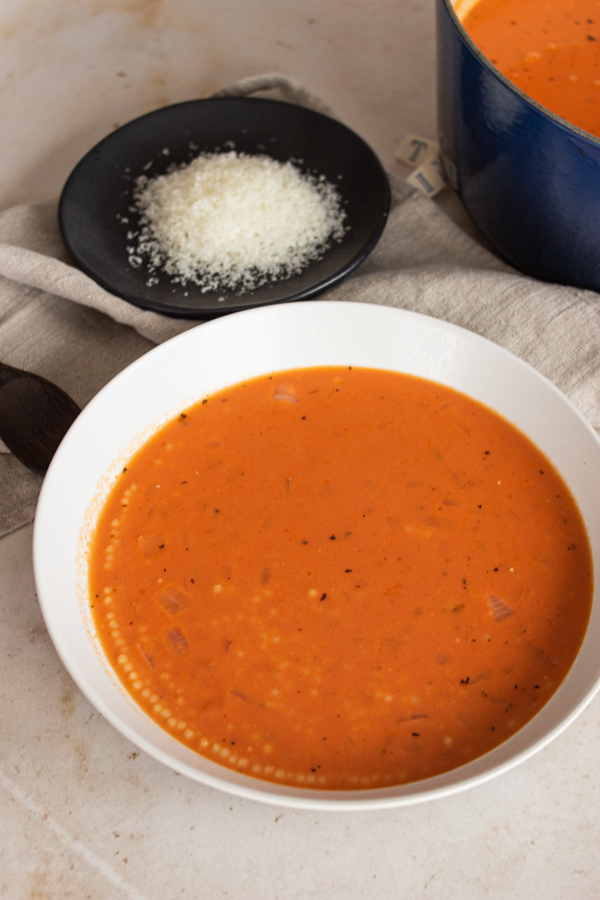 Bowl of tomato pastina soup with grated parmesan and dutch oven in the background. 