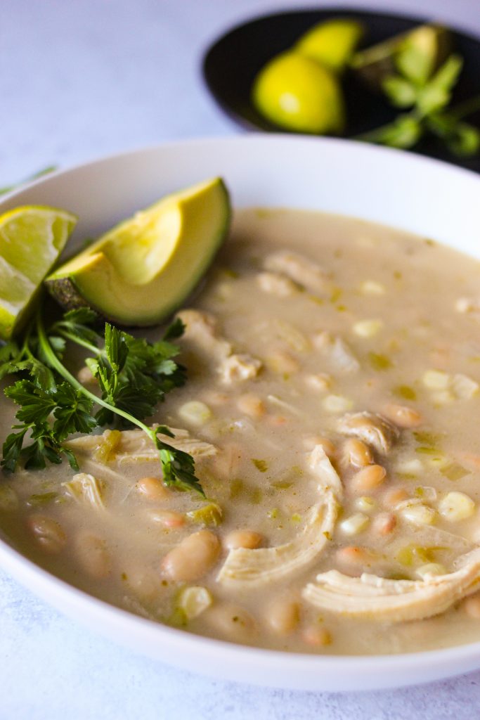 close up view of white chicken chili in a white bowl with avocado and cilantro on the side. 