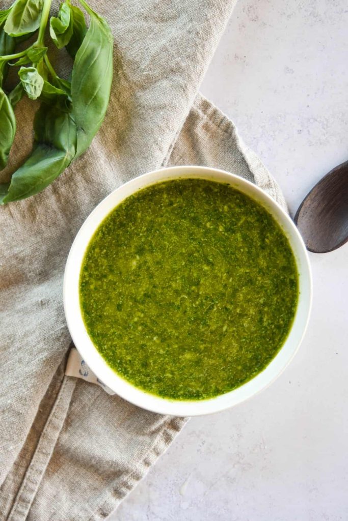 Nut free basil pesto in a small white bowl with a sprig of basil and wooden spoon next to it. 