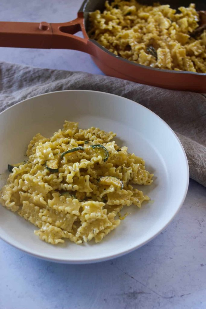 Zucchini pasta in a white bowl with pan of pasta in the background. 