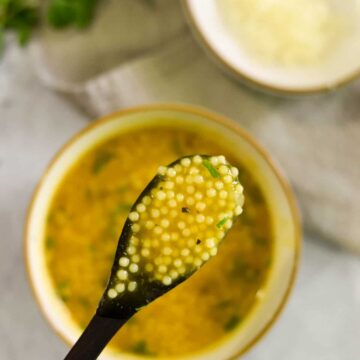 A spoon lifting pastina soup above a bowl.