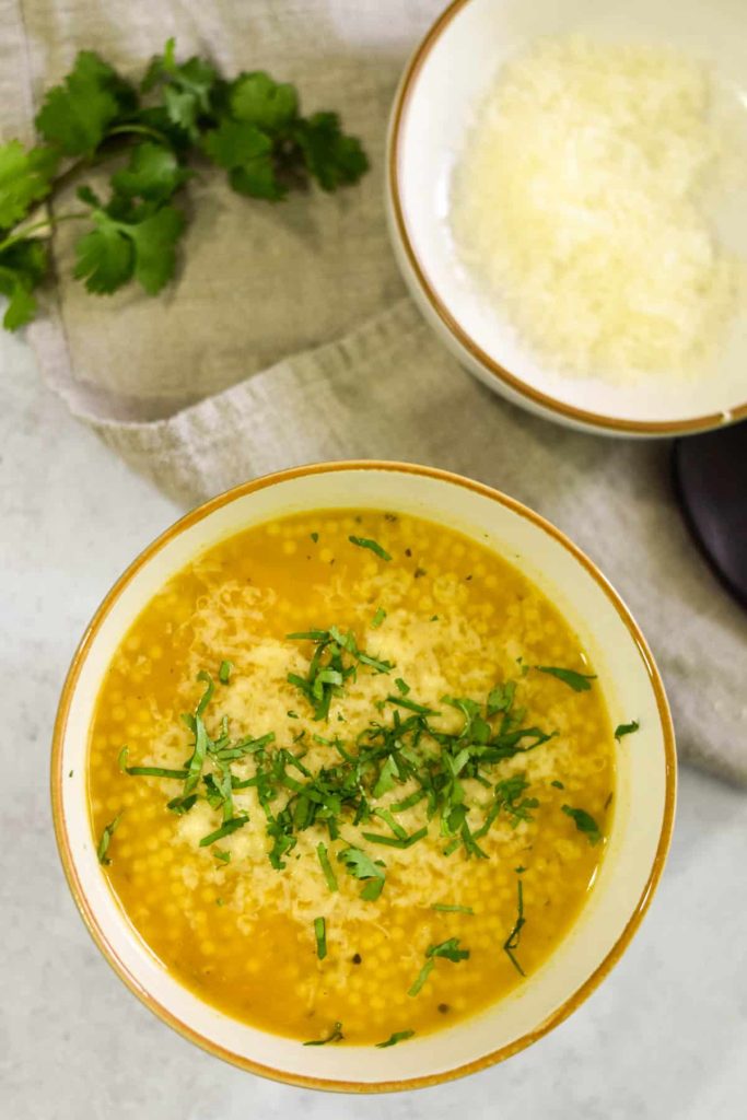 Chicken soup in a bowl with parmesan and parsley on top next to a small bowl with parmesan in it. 