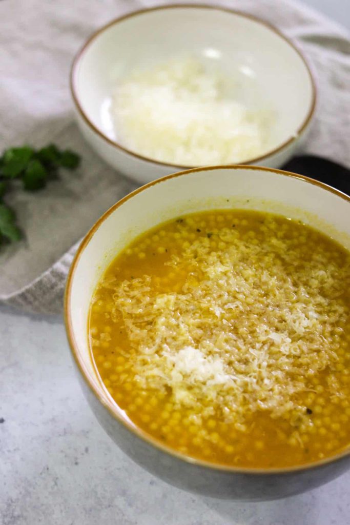 Chicken pastina soup in a bowl with a small bowl of parmesan cheese in the background. 