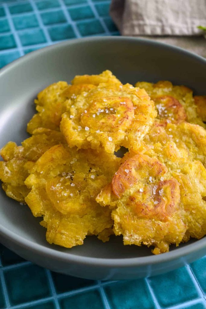 Fried plantains in a shallow grey bowl and topped with crispy salt. 