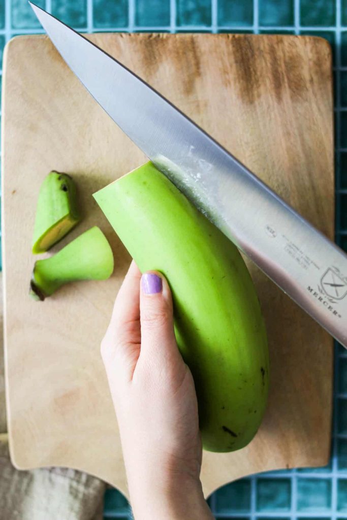 Hand holding a plantain with a chef's knife slicing the edge of the peel. 