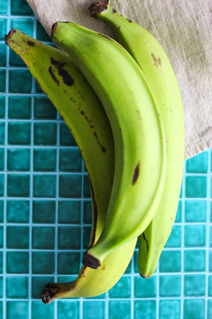 Three green plantains on a blue counter background with a kitchen towel. 