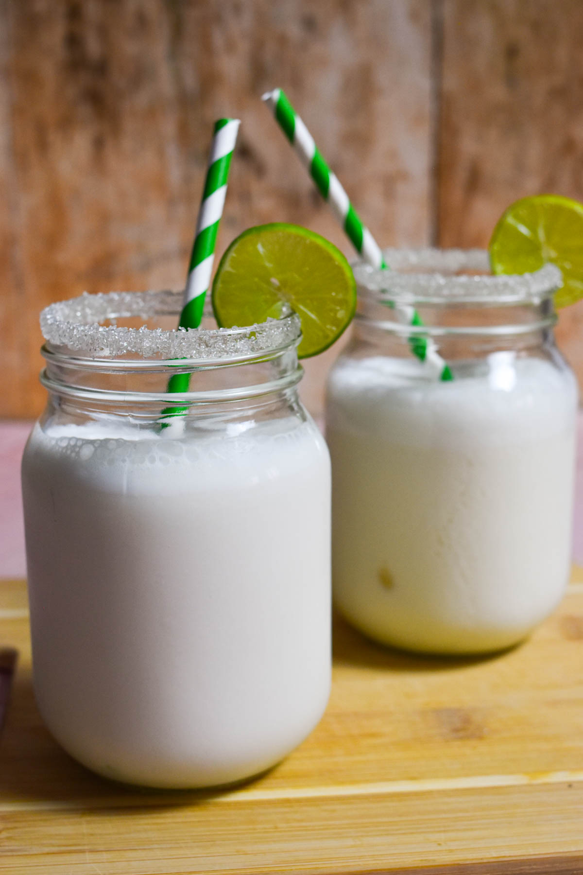 Two jars of limonada de coco with sugar on the rim and a lime wedge, with green and white straws. 