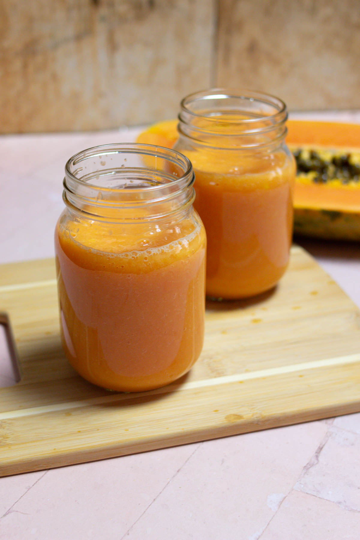 Two papaya mango smoothies in glass jars with a papaya in the background. 