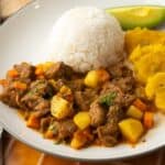 Estofado de carne with potatoes and carrots served with rice and plantains on a plate.