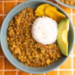 Shallow blue bowl of Ecuadorian lentils with a side of rice, plantains, and sliced avocado.
