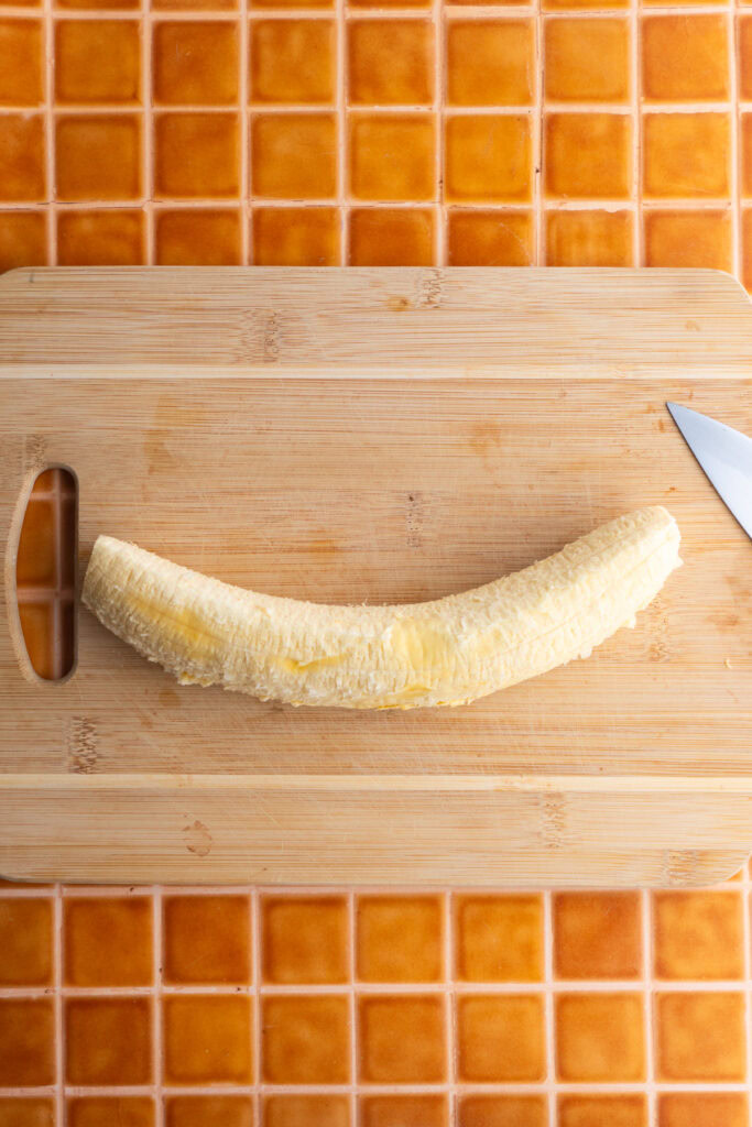 Peeled sweet plantain on a wooden board next to a knife.